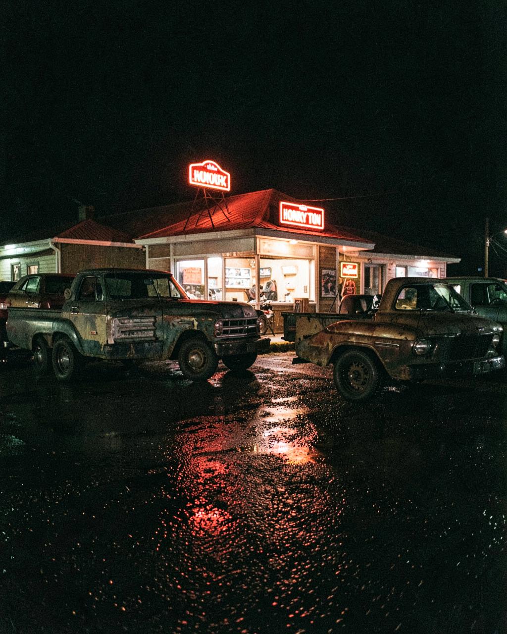Old pickup trucks parked outside a neon-lit small-town bar at night